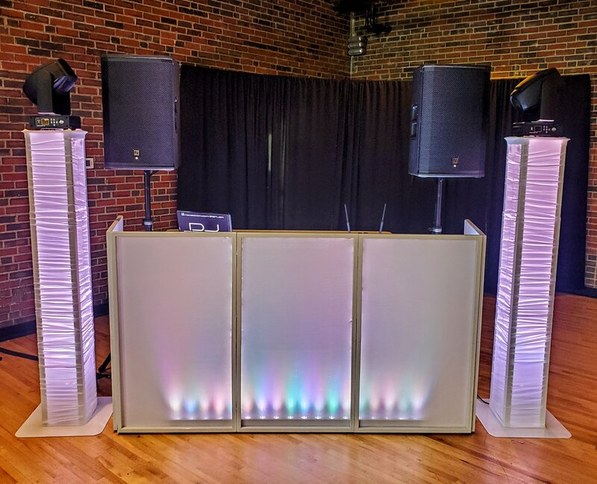 A DJ booth flanked by two illuminated tower speakers and intelligent moving head lights against an exposed brick wall