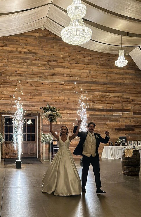 A couple entering their reception with arms raised, framed by two cold spark fountains, in a timber barn hung with chandeliers